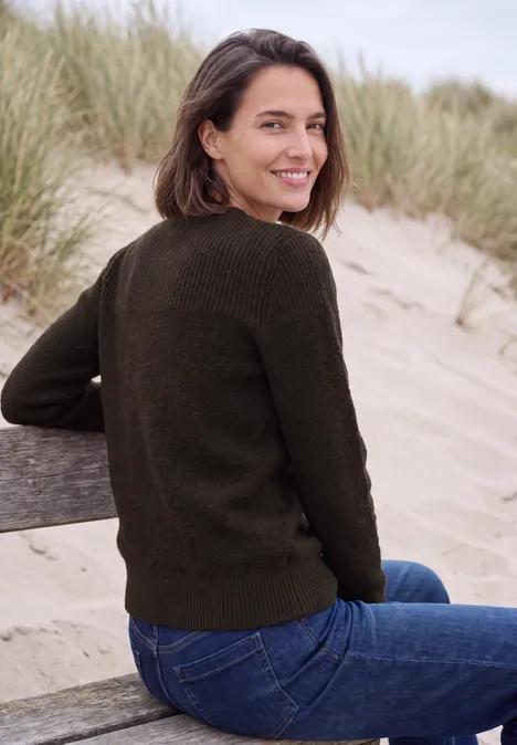 Trui met bouclé-mix en ronde hals truffle brown melange Een glimlachende vrouw in een groen trui en spijkerbroek zit op een bankje aan het strand, duinen op de achtergrond.