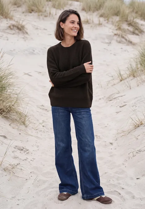 Trui met bouclé-mix en ronde hals truffle brown melange Vrouw lacht op het strand in een donkere trui, wijde blauwe jeans en bruine schoenen voor duinen.