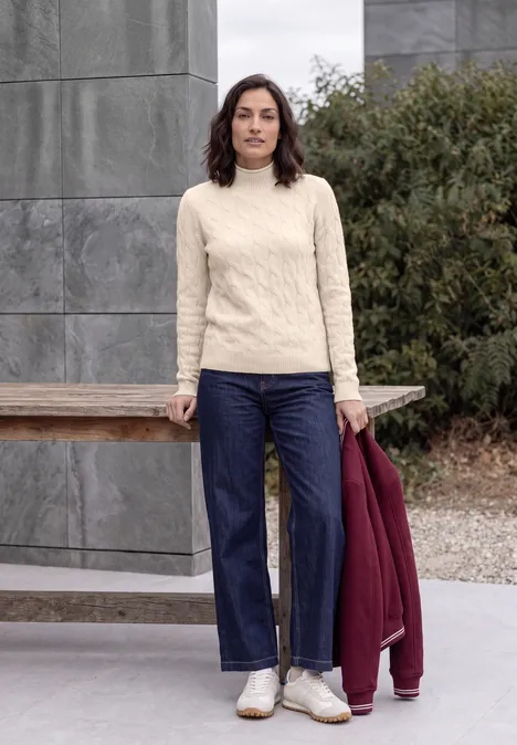 Een vrouw in een beige trui, spijkerbroek en witte schoenen houdt buiten een bordeauxrood jasje vast.