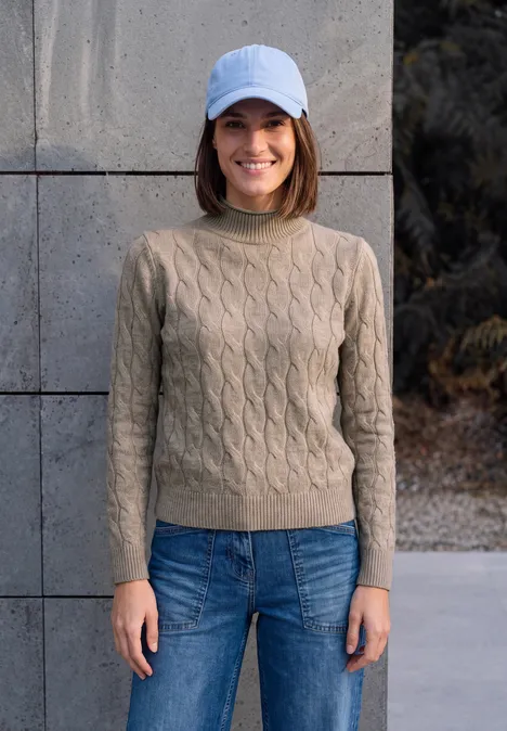 Une femme souriante en pull beige et jeans, coiffée d’une casquette blanche, posant devant un mur gris.