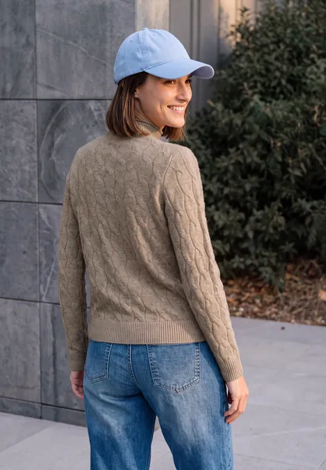 Une jeune femme en jeans, pull marron et casquette bleue regarde et sourit par-dessus son épaule.