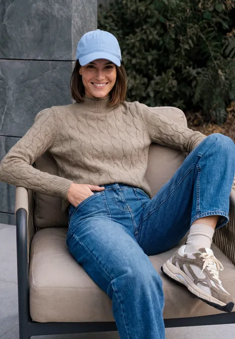 Femme en jean, pull beige et casquette assise de façon décontractée sur une chaise en plein air.