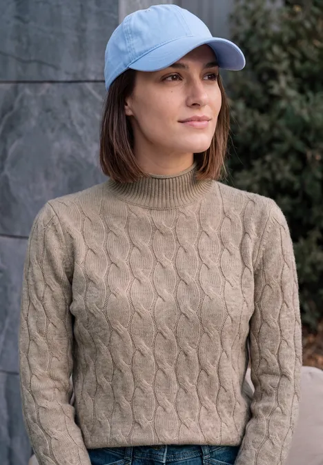Jeune femme portant une casquette bleu clair et un pull torsadé olive, debout devant un mur de pierre à l'extérieur.