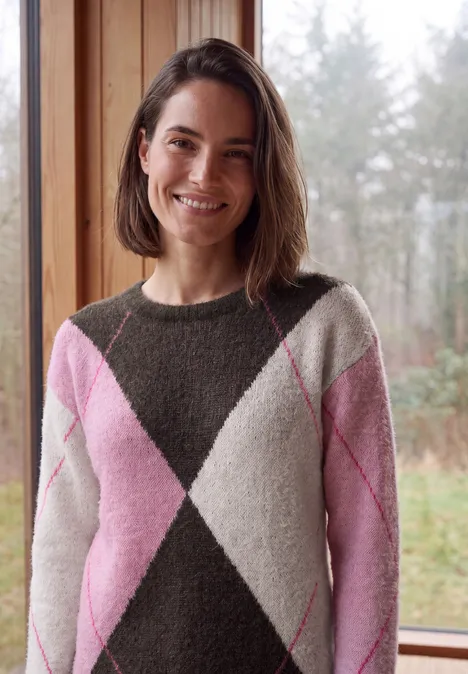 Frau mit schulterlangem Haar im rosa-weißen Rautenmuster-Pullover lächelt vor einem Fenster mit Waldblick.