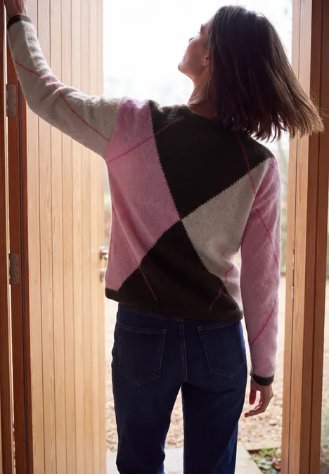 Eine Frau mit Rautenmuster-Pullover in Pink, Grau und Dunkelgrün öffnet von hinten eine Holztür.
