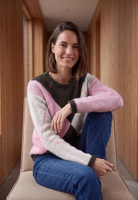Eine Frau sitzt in einem Holzflur, trägt einen bunten Pullover und Jeans, lächelt in die Kamera.