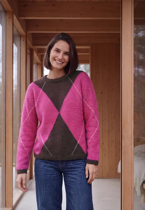 Pullover mit Rundhals und Argyle-Muster pink flash Lächelnde Frau mit rosa-braunem Rautenmuster-Pullover und Jeans steht in einem Holzrahmen und schaut in die Kamera.