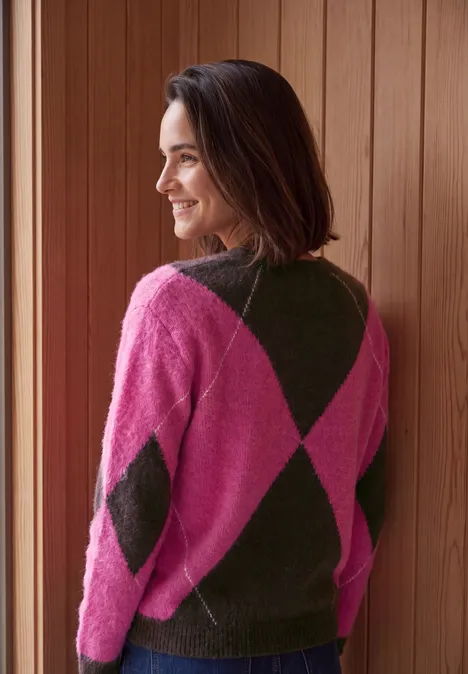Pullover mit Rundhals und Argyle-Muster pink flash Frau lehnt am Fensterrahmen, trägt pink-grünen Rautenmuster-Pullover und lächelt zur Seite.
