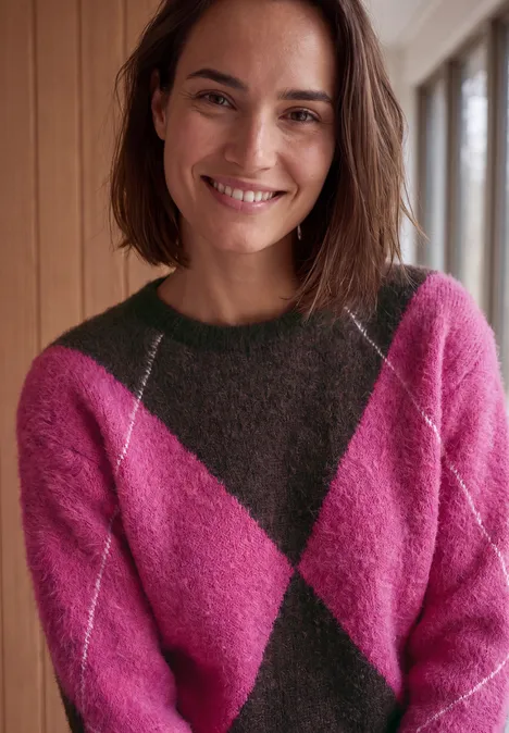 Pullover mit Rundhals und Argyle-Muster pink flash Eine lächelnde Frau mit schulterlangen braunen Haaren trägt einen pink-grün karierten Pullover vor Holzwand.