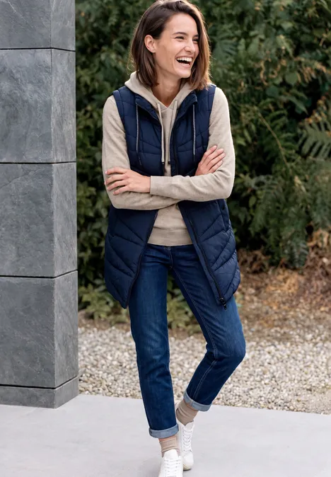 Pull uni à capuche Coffee beige melange Femme souriante en gilet bleu marine, jean et baskets blanches, appuyée sur une colonne dans un jardin.