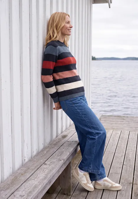Eine junge Frau lehnt lässig an einer weißen Wand auf einem Steg, trägt Pullover, Jeans und Sneaker am Wasser.