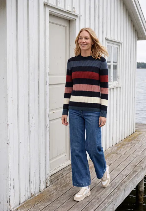 Eine lächelnde Frau in Jeans und gestreiftem Pullover läuft auf einem Steg vor einem weißen Haus am Wasser.