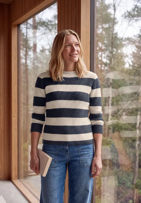 Pullover mit Turtle Neck und Streifen urban dark blue Eine Frau mit gestreiftem Pullover hält ein Buch und steht vor einem Fenster mit Waldsicht.
