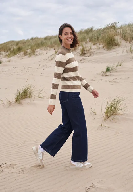 Pullover mit Turtle Neck und Streifen creamy mocha melange Frau spaziert lächelnd am Sandstrand in gestreiftem Pullover, weiten Jeans und weißen Sneakern.