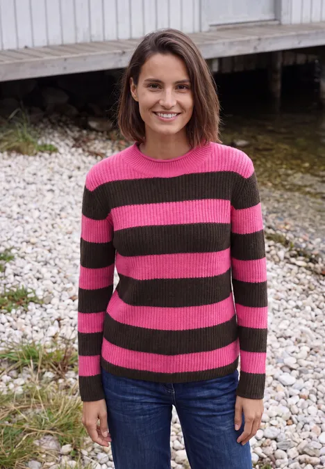 Pullover mit Turtle Neck und Streifen pink flash Frau mit rosa-grün gestreiftem Pullover und Jeans steht lächelnd am Ufer vor einem Holzsteg.