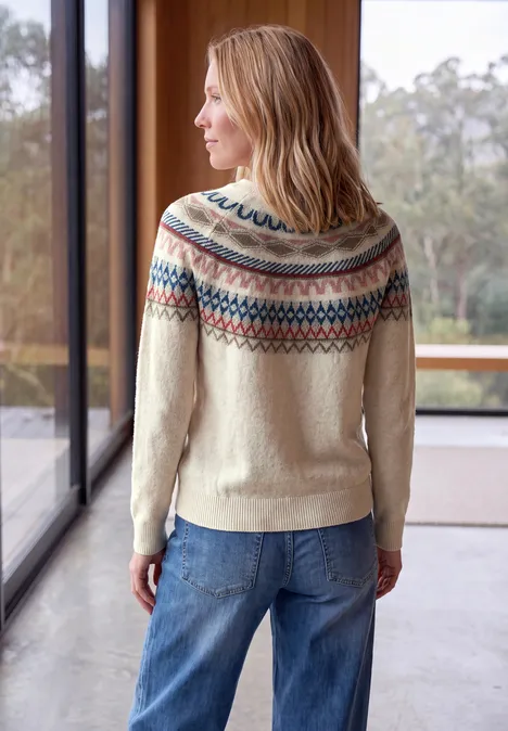 Pull jacquard à col rond oat milk beige melange Femme de dos portant un pull Fair Isle clair et un jean, devant de grandes fenêtres dans une pièce lumineuse.