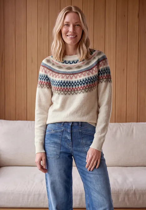 Pull jacquard à col rond oat milk beige melange Femme souriante en pull jacquard et jean, devant un mur en bois et un canapé.