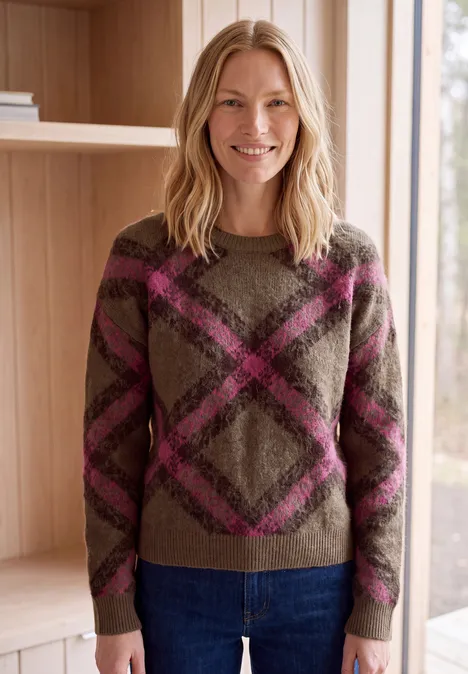 Eine lächelnde Frau in einem braun-rosa Rautenmuster-Pullover und blauer Jeans in einem hellen Holzraum.