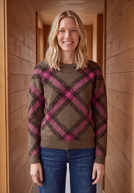 Eine Frau steht in einem Holzflur, trägt einen karierten Pullover und Jeans und lächelt in die Kamera.