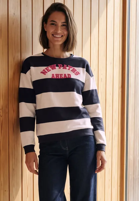 Frau mit dunkelblau-weiß gestreiftem Pullover und Jeans steht lächelnd vor einer Holzvertäfelung.