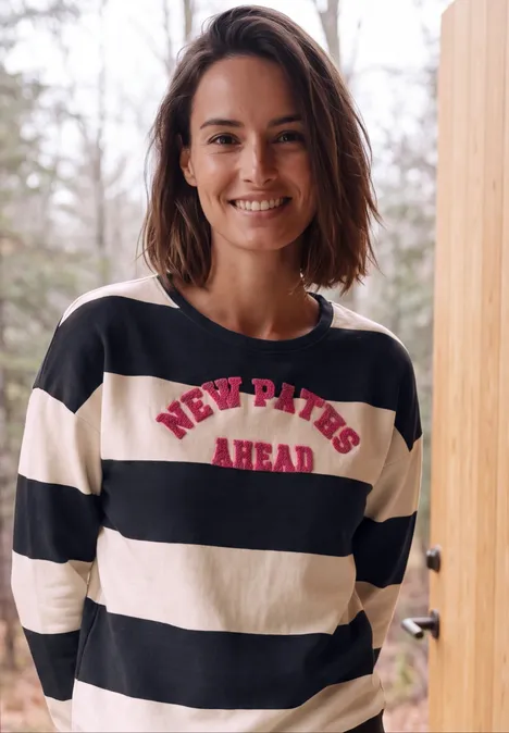 Lächelnde Frau in schwarz-weiß gestreiftem Shirt mit rotem Schriftzug NEW PATHS AHEAD vor Holzbalken im Freien.