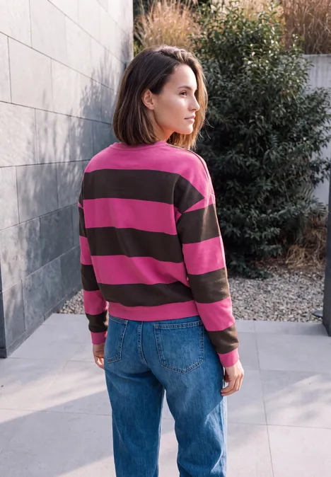 Eine Frau steht im Freien mit pink-braun gestreiftem Pullover und Jeans vor einer grauen Wand und Büschen.