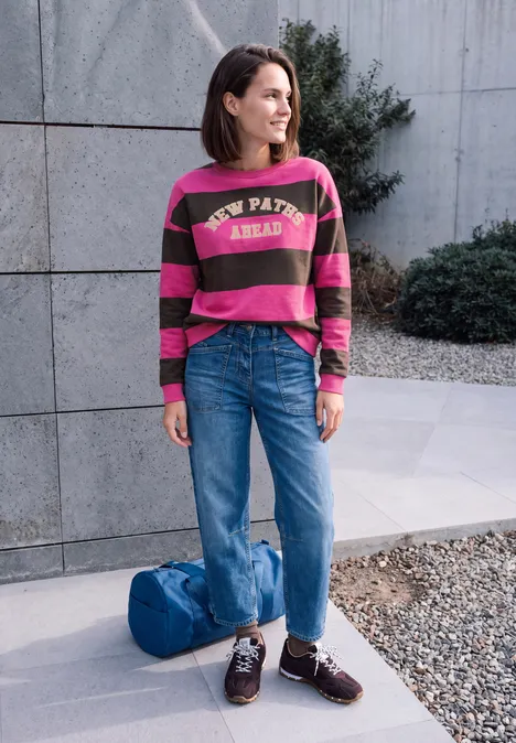 Eine Frau vor einer Betonwand, trägt gestreiftes Sweatshirt, weite Jeans und Lederschuhe, Tasche am Boden.