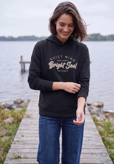 Sweatshirt mit Details in Rippstruktur urban dark blue Eine lächelnde Frau in Jeans und schwarzem Kapuzenpullover steht auf einem Steg am Wasser.