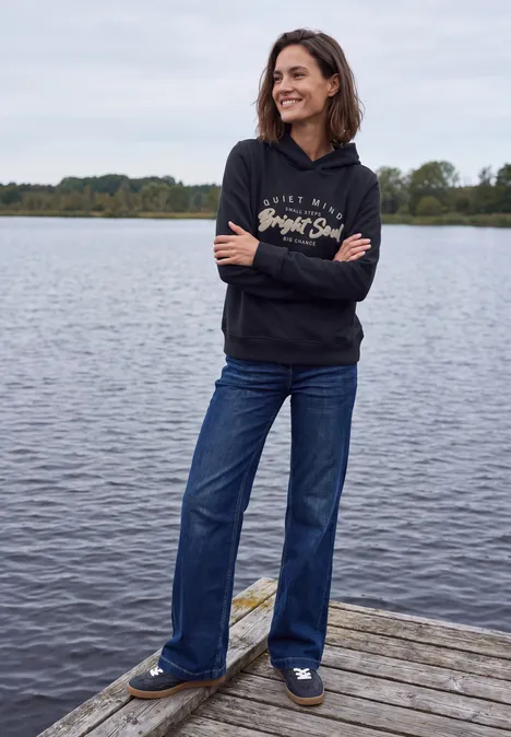 Sweatshirt mit Details in Rippstruktur urban dark blue Eine Frau in schwarzer Kapuzenjacke und blauen Jeans steht lächelnd auf einem Holzsteg am Wasser.