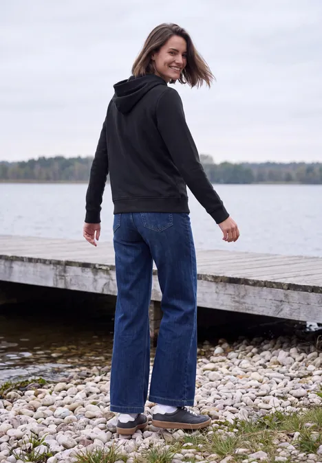 Sweatshirt mit Details in Rippstruktur urban dark blue Eine Frau in schwarzer Kapuzenjacke und blauen Jeans steht am Ufer, blickt lächelnd zurück.