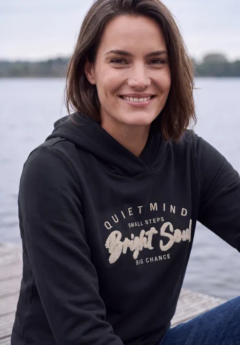 Sweatshirt mit Details in Rippstruktur urban dark blue Eine lächelnde Frau in schwarzem Hoodie sitzt entspannt an einem Seeufer.