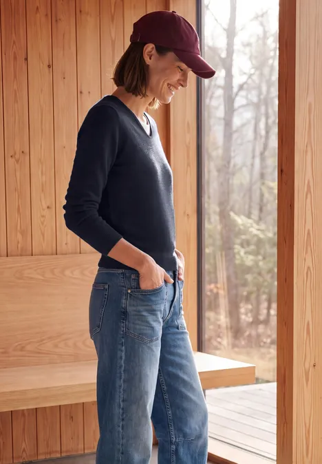 Frau in Jeans, dunkelblauem Pullover und Cap steht lächelnd seitlich vor einer Holzhütte im Wald.