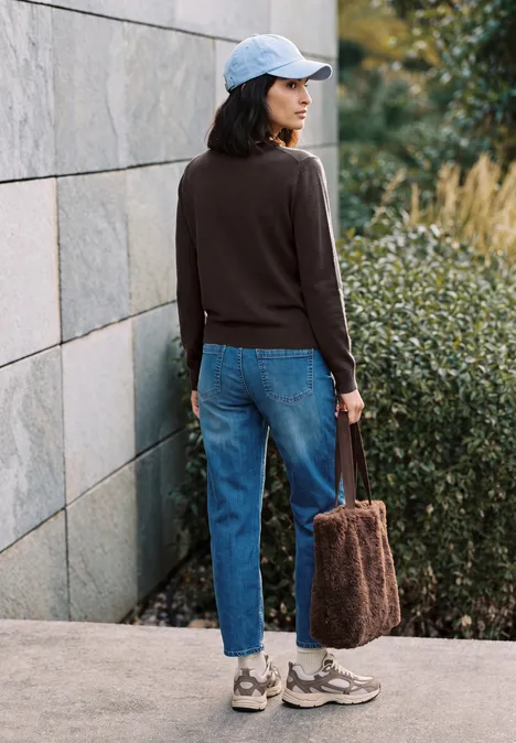 Femme en jean, pull marron foncé et casquette, sac en peluche, debout près d’un mur et de buissons verts.