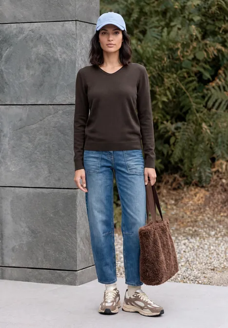 Femme en casquette bleue, pull marron, jean, baskets et sac en fausse fourrure devant un mur végétal.
