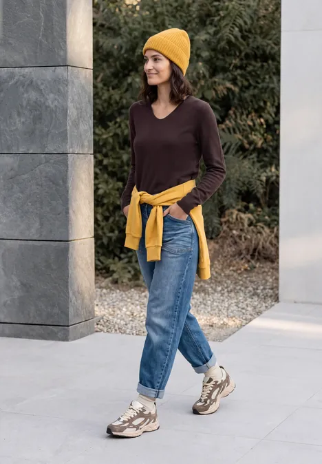 Pullover mit gerundetem V-Ausschnitt berry red Frau in gelber Mütze, braunem Pullover, Jeans und Sneakers spaziert lächelnd im Freien.