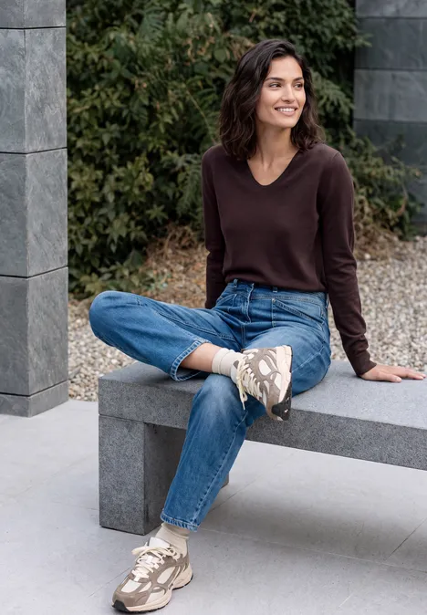Pullover mit gerundetem V-Ausschnitt berry red Eine lächelnde Frau sitzt im Freien auf einer Steinbank, trägt braunen Pullover, blaue Jeans und beige Sneaker.