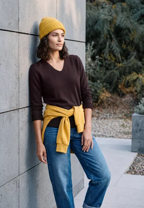 Pullover mit gerundetem V-Ausschnitt berry red Frau lehnt lässig an Betonwand, trägt braunen Pullover, Jeans, gelbe Mütze und Sweater um die Hüfte.