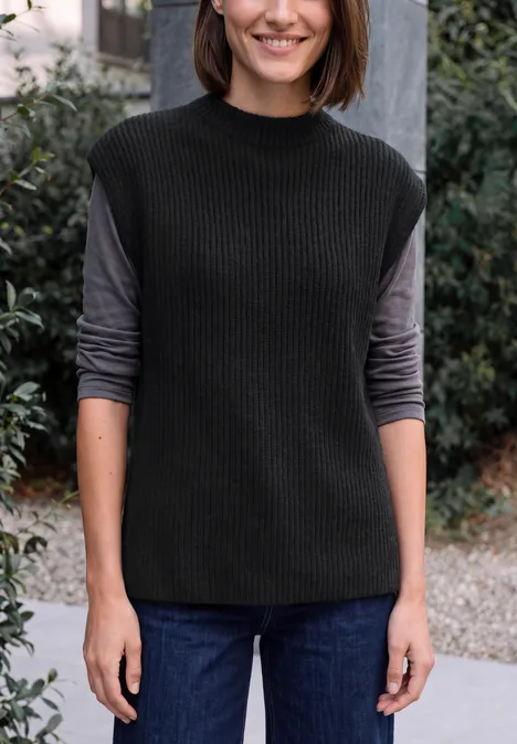 Pullunder mit Turtle Neck und Zopfdetails Black Frau mit schwarzer Strickweste über grauem Langarmshirt und Jeans, stehend vor einem Büschehintergrund.