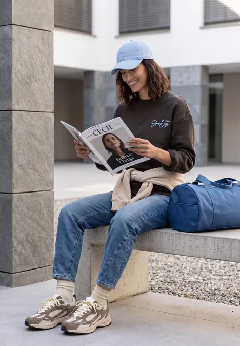 Eine Frau in Basecap liest entspannt eine Zeitschrift an einem Gebäude neben ihrer blauen Reisetasche.