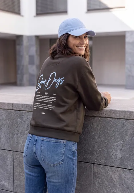 Eine Frau in Jeans, dunkelgrünem Sweatshirt mit Aufdruck und hellblauer Cap lehnt lächelnd an einer Steinmauer.