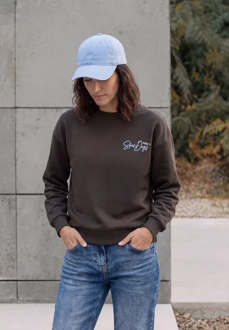 Frau in weißer Baseballkappe, braunem Sweatshirt und Jeans, die im Freien nach unten schaut.