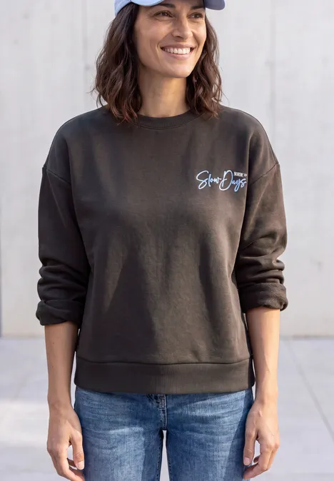 Junge Frau in Jeans, dunklem Pullover mit Schriftzug und blauer Baseballkappe, steht lächelnd vor hellem Hintergrund.