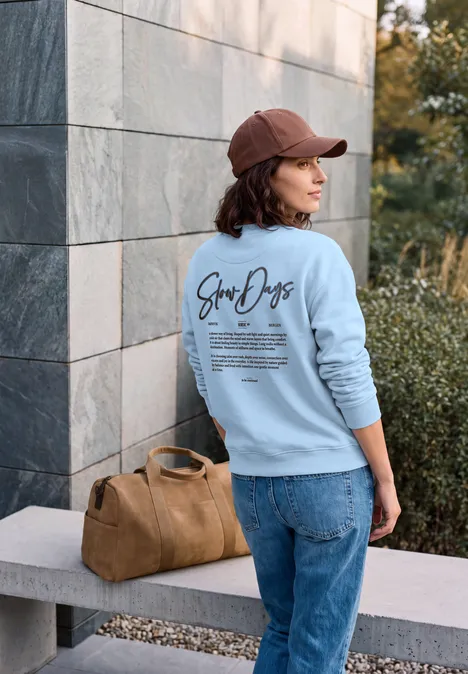 Frau von hinten in hellblauem Sweatshirt und roter Kappe steht an einer Steinbank mit brauner Tasche.