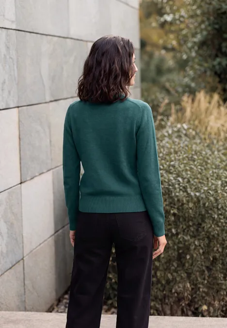 Pullover in Unifarbe mit Turtle Neck dark sea green melange Frau in grünem Pullover und schwarzer Hose, Rücken zur Kamera, steht vor einer Steinmauer mit Büschen.