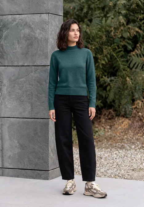 Pullover in Unifarbe mit Turtle Neck dark sea green melange Frau in grünem Pullover, schwarzen Hosen und Sneakers steht vor einer grauen Betonwand im Freien.