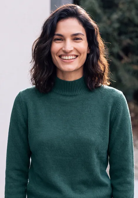 Pullover in Unifarbe mit Turtle Neck dark sea green melange Eine lächelnde Frau mit dunklen Haaren und grünem Pullover vor unscharfem Hintergrund im Freien.