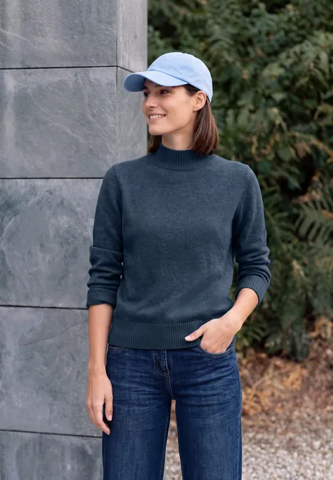 Pullover in Unifarbe mit Turtle Neck cotton blue melange Frau in dunkelblauem Pullover, Jeans und Cap steht lächelnd vor Steinwand und grünen Büschen.