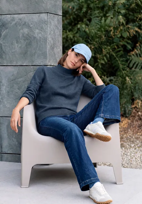 Pullover in Unifarbe mit Turtle Neck cotton blue melange Frau sitzt entspannt draußen in einem beigen Stuhl, trägt Jeans, Pullover, weiße Sneaker und blaue Cap.