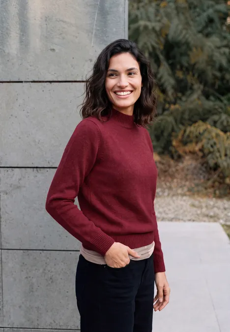 Pullover in Unifarbe mit Turtle Neck rhubarb melange Eine lächelnde Frau mit braunen Haaren und rotem Pullover steht vor einer grauen Wand im Freien.