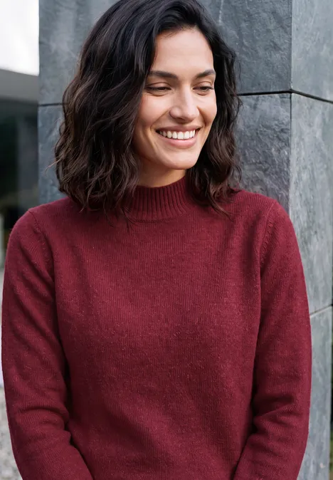Pullover in Unifarbe mit Turtle Neck rhubarb melange Lächelnde Frau in rotem Pullover lehnt an einer grauen Wand.